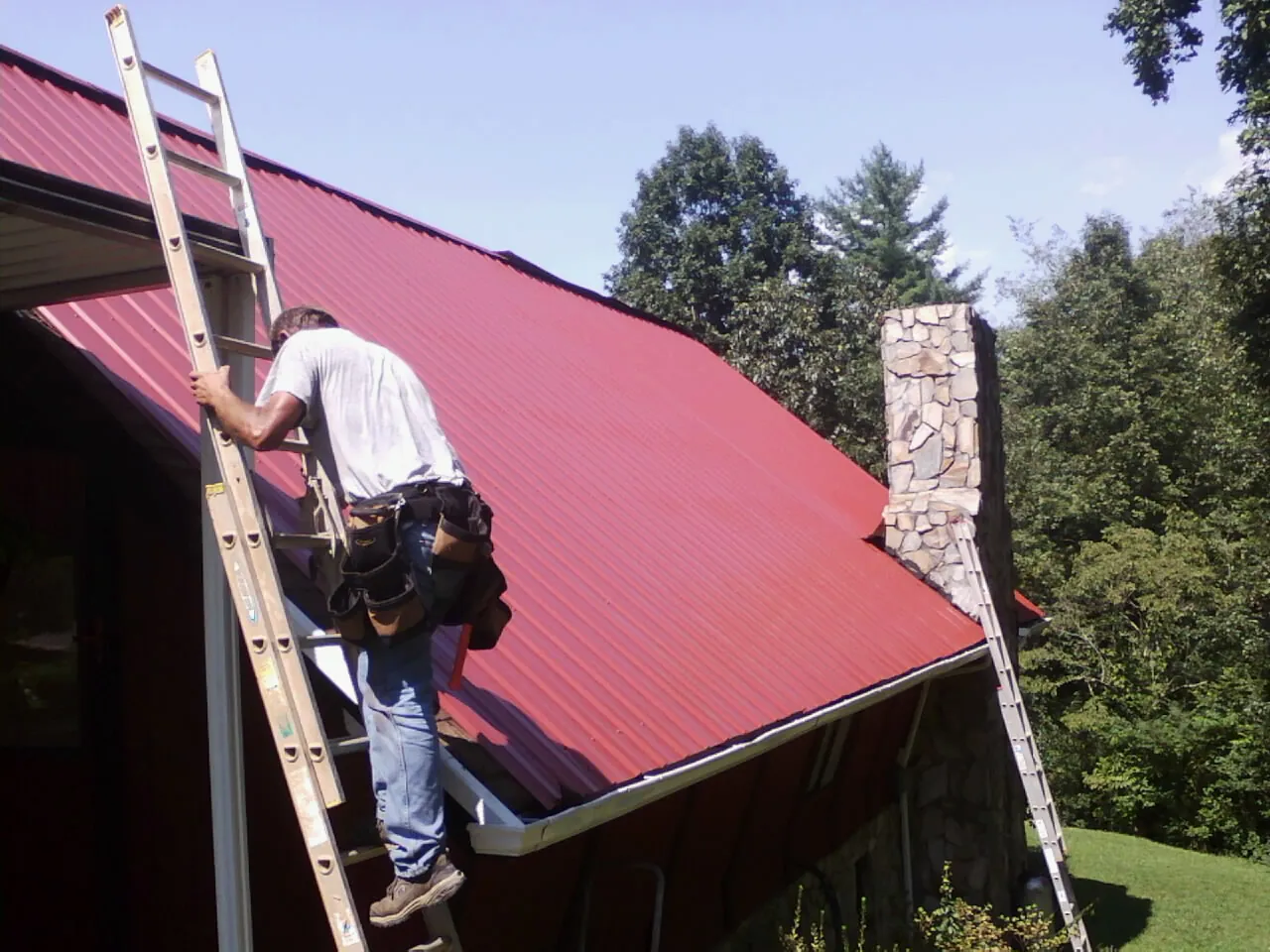 Roofer on a ladder beside a red metal roof and stone chimney — Soffit & Fascia Repair by Roofing Brothers in Vandalia